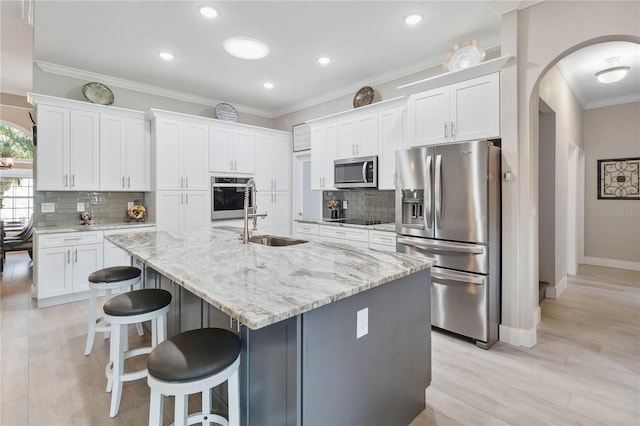 kitchen with light stone counters, a center island with sink, ornamental molding, appliances with stainless steel finishes, and white cabinets