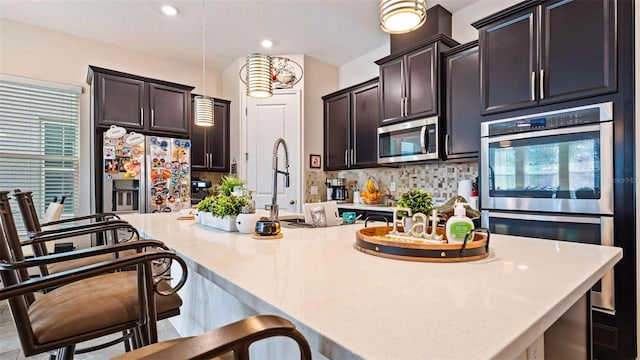 kitchen with tasteful backsplash, decorative light fixtures, dark brown cabinets, appliances with stainless steel finishes, and an island with sink