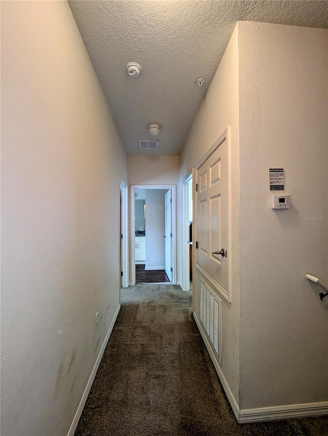 hallway with dark colored carpet, visible vents, a textured wall, a textured ceiling, and baseboards