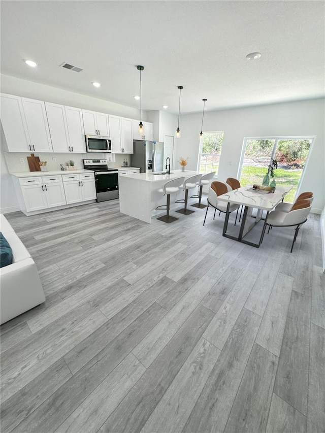 kitchen with pendant lighting, light hardwood / wood-style flooring, appliances with stainless steel finishes, white cabinets, and a center island with sink