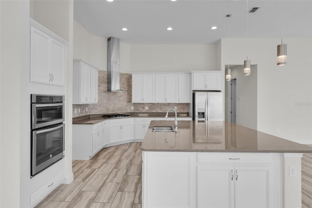 kitchen with wall chimney exhaust hood, sink, white cabinetry, appliances with stainless steel finishes, and a kitchen island with sink