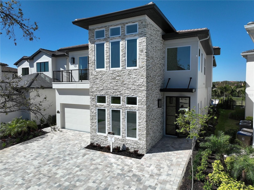 view of front facade featuring a balcony, a garage, stone siding, decorative driveway, and stucco siding