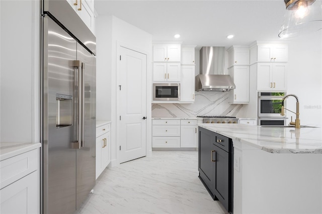 kitchen with white cabinets, built in appliances, light stone countertops, and wall chimney range hood