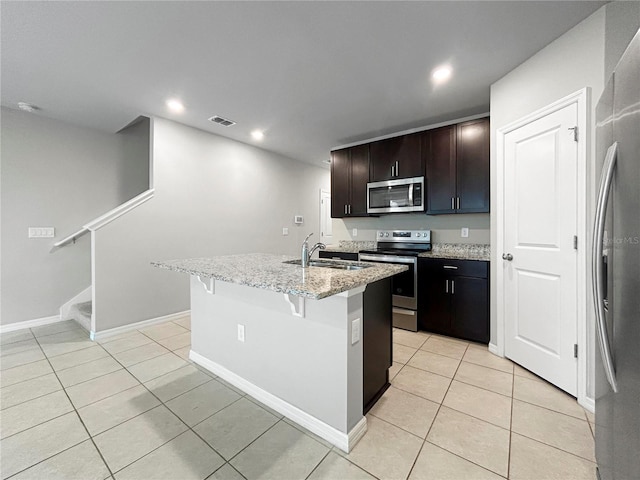 kitchen with dark brown cabinetry, light tile patterned floors, appliances with stainless steel finishes, an island with sink, and light stone countertops