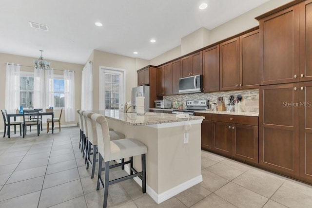 kitchen with appliances with stainless steel finishes, a kitchen island with sink, a kitchen breakfast bar, light stone counters, and tasteful backsplash