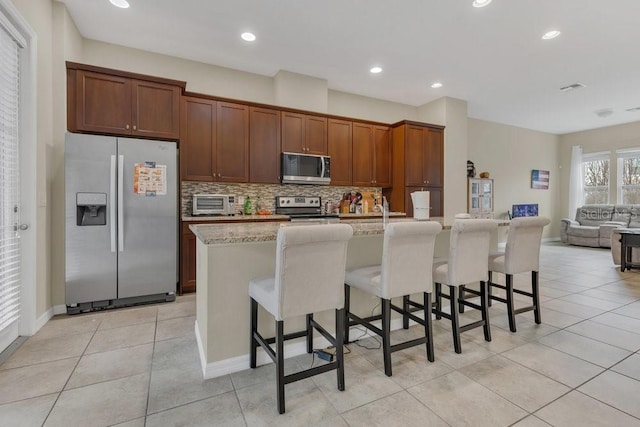 kitchen featuring appliances with stainless steel finishes, light stone counters, an island with sink, a kitchen bar, and decorative backsplash