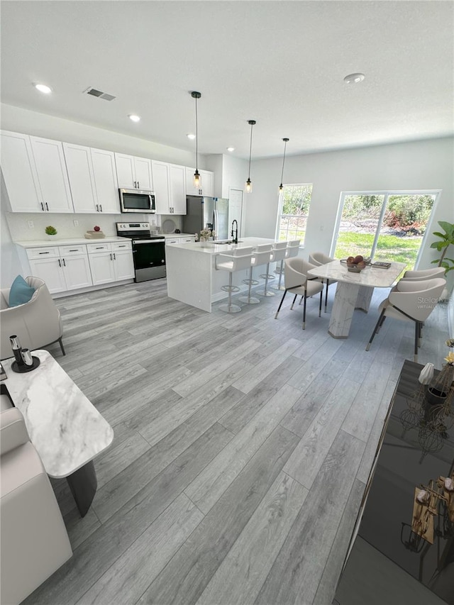 kitchen with appliances with stainless steel finishes, white cabinetry, hanging light fixtures, a center island with sink, and light wood-type flooring