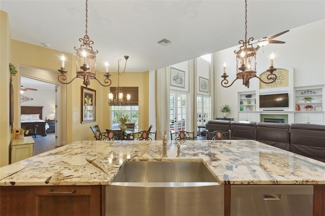 kitchen featuring hanging light fixtures, ceiling fan with notable chandelier, a center island with sink, and light stone counters