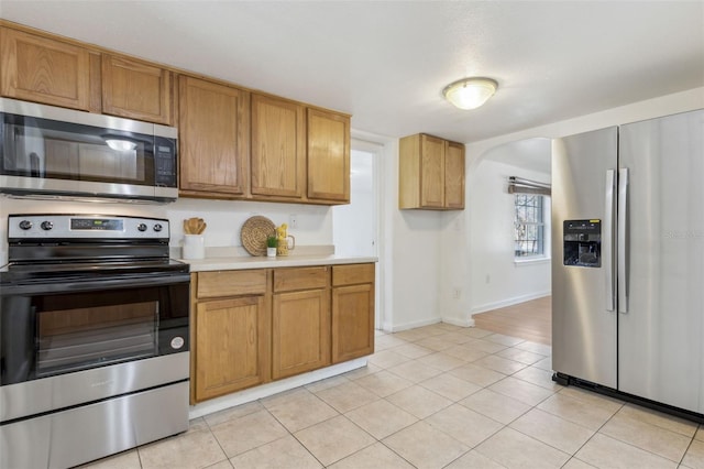 kitchen featuring arched walkways, brown cabinets, stainless steel appliances, light countertops, and light tile patterned flooring