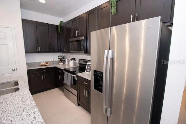 kitchen featuring light tile patterned floors, appliances with stainless steel finishes, dark brown cabinetry, a sink, and light stone countertops