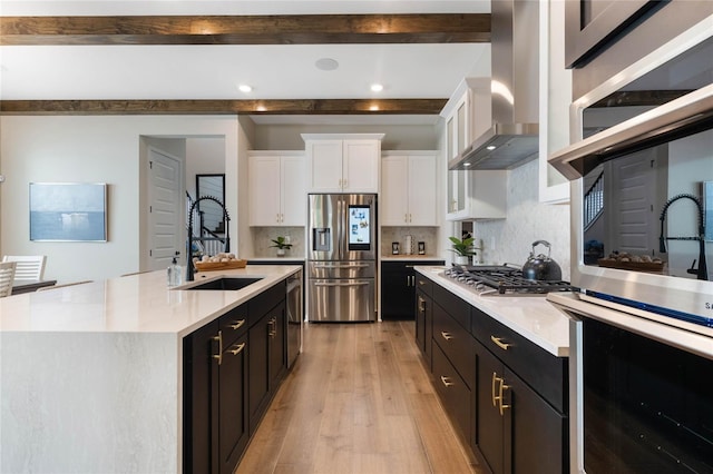 kitchen with wall chimney range hood, sink, white cabinetry, stainless steel appliances, and a center island with sink