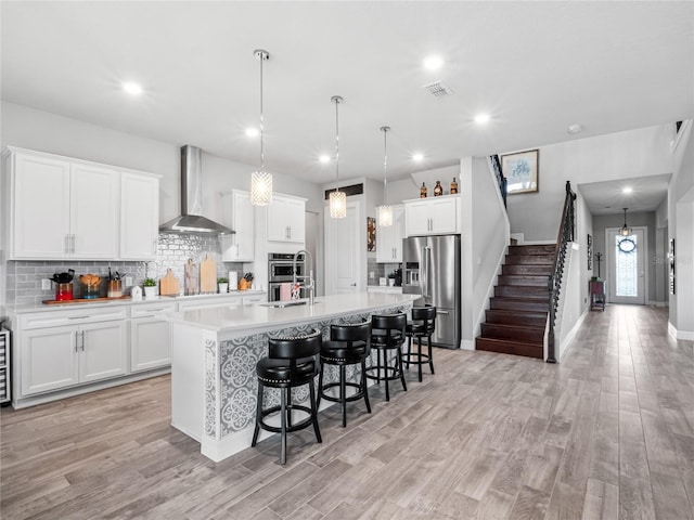 kitchen featuring wall chimney exhaust hood, a kitchen island with sink, appliances with stainless steel finishes, and light countertops