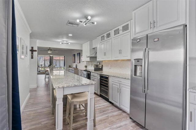 kitchen featuring wine cooler, white cabinets, stainless steel appliances, light stone countertops, and wall chimney range hood