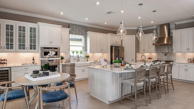 kitchen with wall chimney range hood, appliances with stainless steel finishes, hanging light fixtures, white cabinets, and a center island with sink