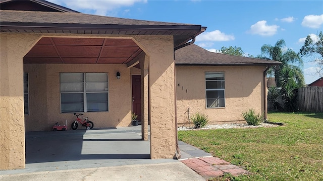 doorway to property featuring a shingled roof, a lawn, fence, and stucco siding