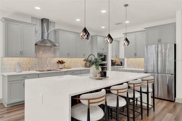 kitchen with gray cabinets, stainless steel fridge, hanging light fixtures, wall chimney range hood, and a center island with sink