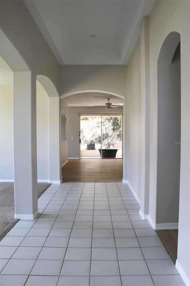 hallway with arched walkways, a raised ceiling, baseboards, and light tile patterned floors