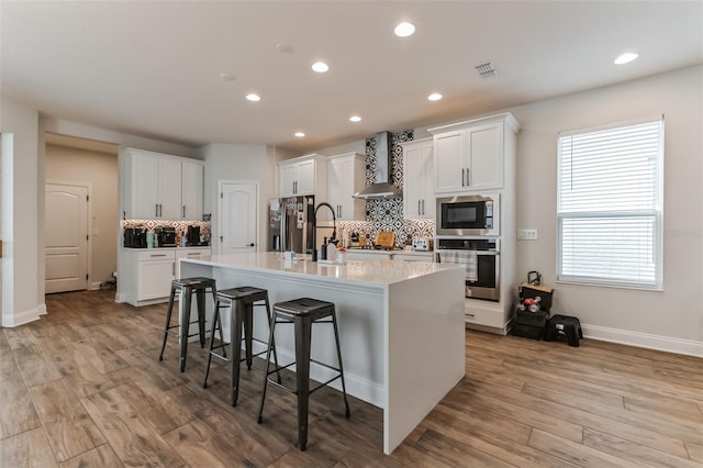 kitchen with wall chimney range hood, a breakfast bar, appliances with stainless steel finishes, an island with sink, and white cabinets