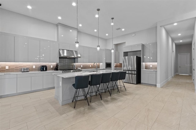 kitchen with a breakfast bar, freestanding refrigerator, under cabinet range hood, a towering ceiling, and modern cabinets