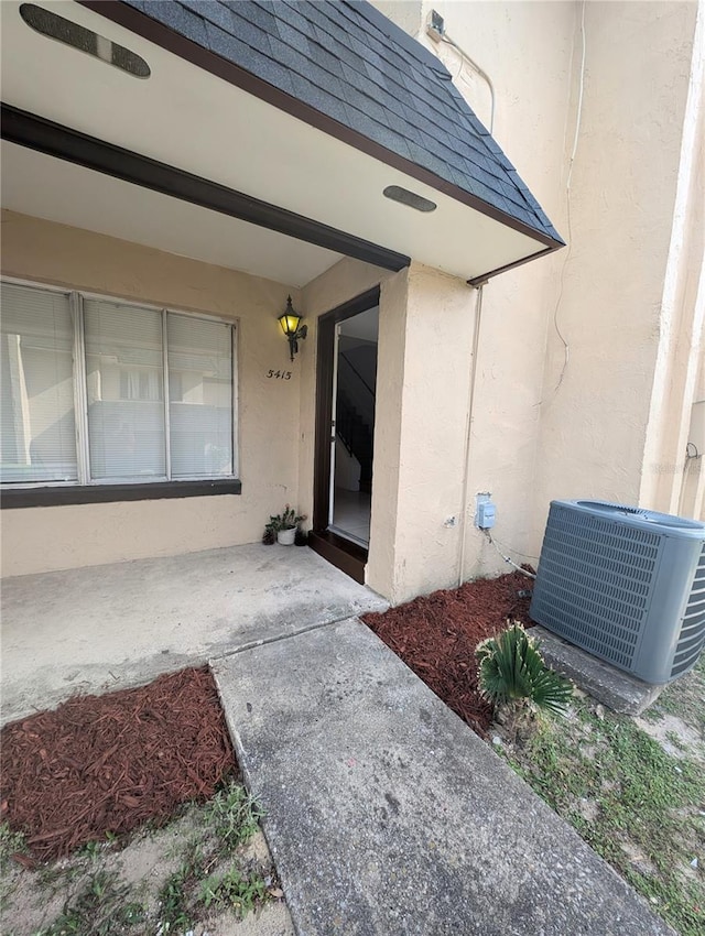 view of exterior entry with a shingled roof, cooling unit, a patio area, and stucco siding