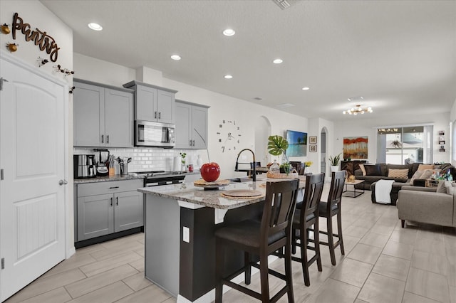 kitchen with a kitchen bar, light stone counters, appliances with stainless steel finishes, gray cabinets, and an island with sink