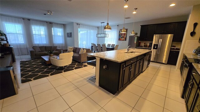 kitchen featuring sink, light stone counters, stainless steel fridge with ice dispenser, hanging light fixtures, and a center island with sink