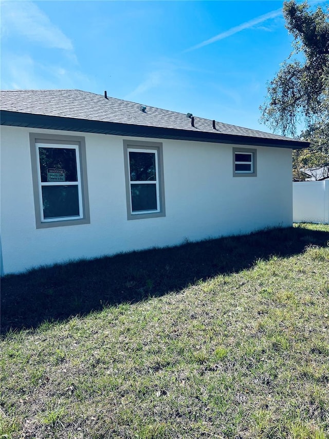 view of side of property featuring roof with shingles, a lawn, fence, and stucco siding