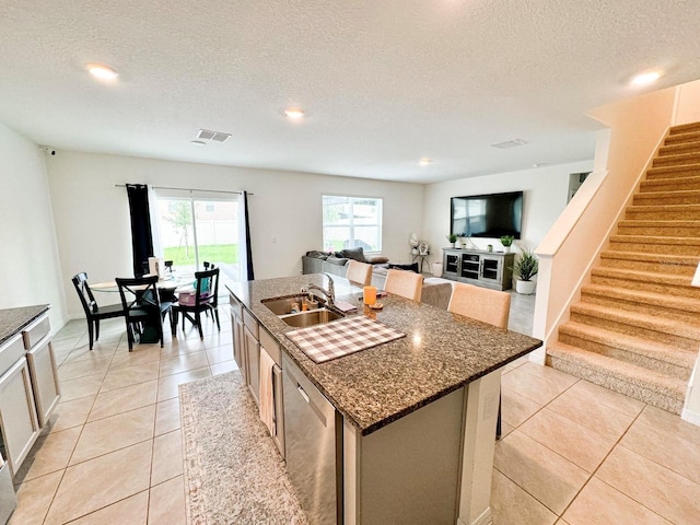 kitchen featuring sink, light tile patterned floors, dishwasher, a kitchen island with sink, and dark stone counters