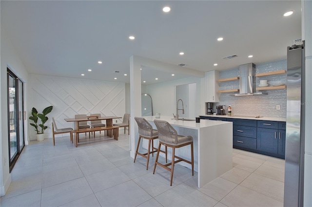 kitchen with blue cabinets, a breakfast bar area, a center island with sink, decorative backsplash, and wall chimney range hood