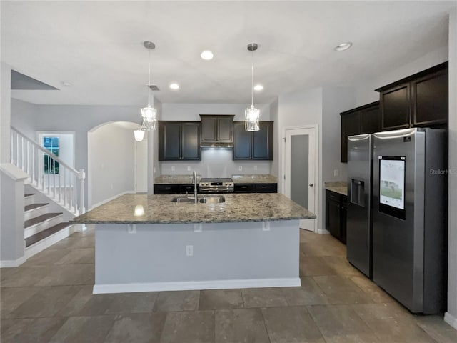 kitchen featuring sink, hanging light fixtures, stainless steel fridge with ice dispenser, light stone countertops, and a center island with sink