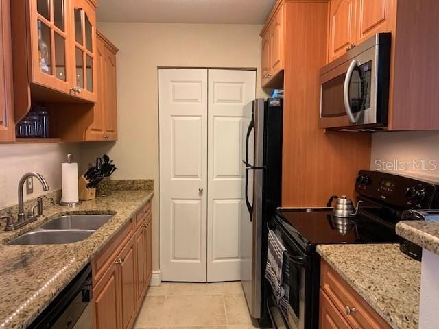 kitchen with brown cabinetry, glass insert cabinets, appliances with stainless steel finishes, light stone countertops, and a sink