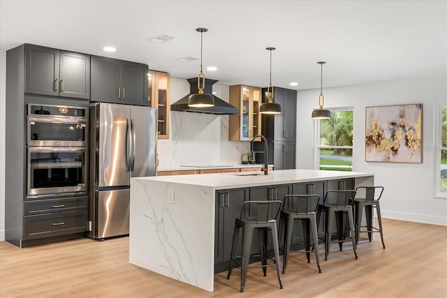 kitchen with appliances with stainless steel finishes, wall chimney range hood, a kitchen island with sink, and light wood-style floors