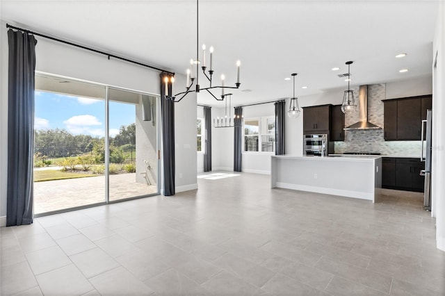 kitchen featuring backsplash, hanging light fixtures, a kitchen island with sink, dark brown cabinetry, and wall chimney range hood