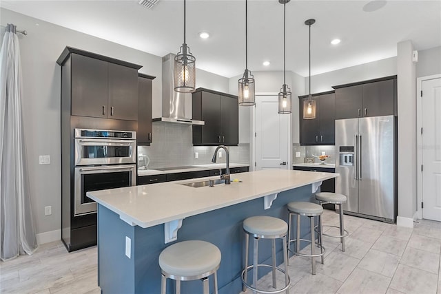 kitchen featuring an island with sink, wall chimney exhaust hood, appliances with stainless steel finishes, light countertops, and pendant lighting