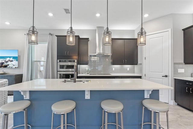 kitchen with light countertops, stainless steel double oven, an island with sink, wall chimney exhaust hood, and black electric cooktop