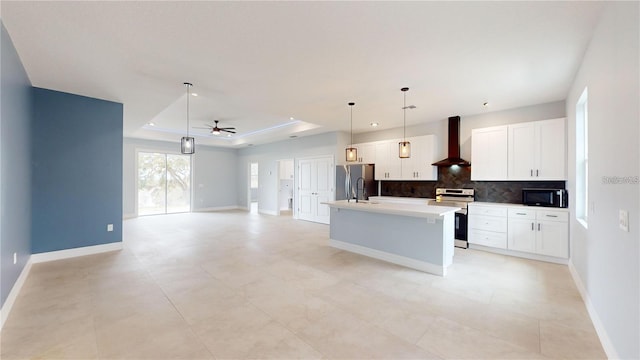 kitchen with appliances with stainless steel finishes, white cabinetry, a kitchen island with sink, a raised ceiling, and wall chimney exhaust hood