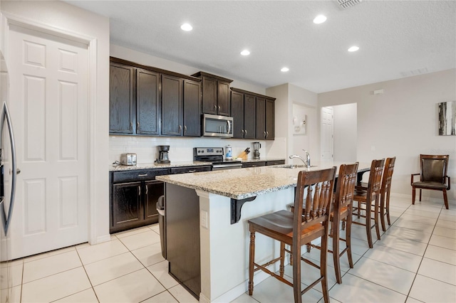 kitchen with a breakfast bar, appliances with stainless steel finishes, a kitchen island with sink, tasteful backsplash, and light stone countertops