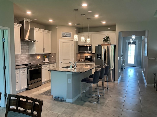 kitchen with a center island with sink, appliances with stainless steel finishes, white cabinets, a sink, and wall chimney exhaust hood