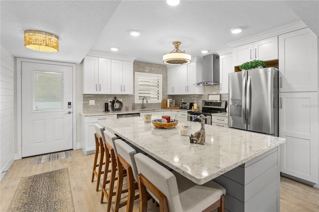 kitchen featuring light wood finished floors, appliances with stainless steel finishes, white cabinetry, wall chimney exhaust hood, and a sink