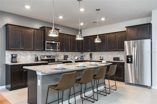 kitchen featuring appliances with stainless steel finishes, decorative light fixtures, a kitchen island with sink, dark brown cabinetry, and light stone countertops