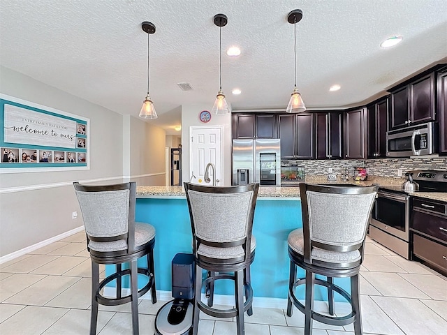 kitchen featuring dark brown cabinetry, decorative light fixtures, appliances with stainless steel finishes, an island with sink, and decorative backsplash