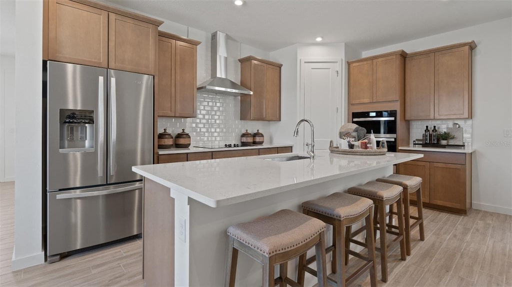 kitchen featuring sink, a kitchen island with sink, stainless steel refrigerator with ice dispenser, a kitchen bar, and wall chimney exhaust hood