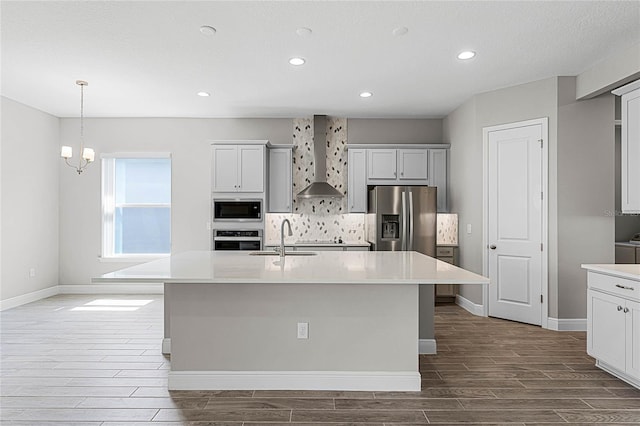 kitchen with wall chimney range hood, sink, appliances with stainless steel finishes, a kitchen island with sink, and decorative backsplash