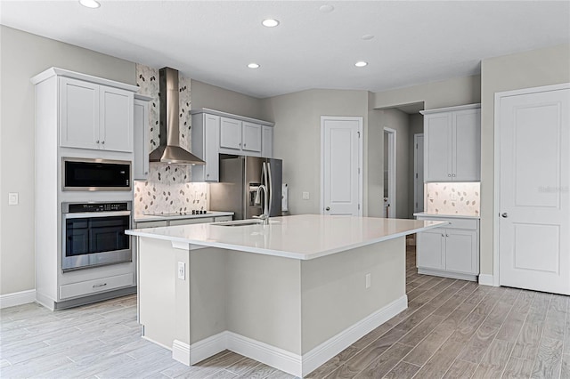 kitchen featuring appliances with stainless steel finishes, a kitchen island with sink, light wood-type flooring, and wall chimney exhaust hood