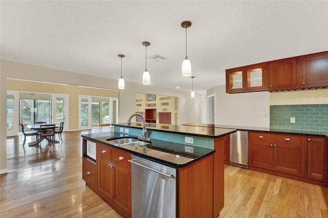 kitchen with a center island with sink, dark stone counters, stainless steel dishwasher, pendant lighting, and a sink