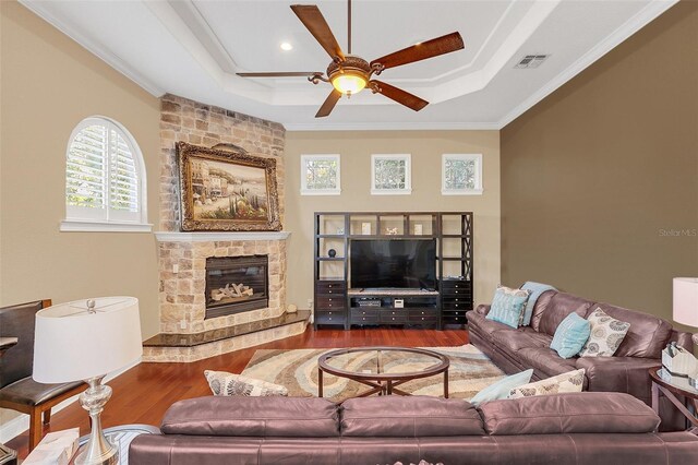 living area with ceiling fan, a stone fireplace, wood finished floors, a tray ceiling, and crown molding