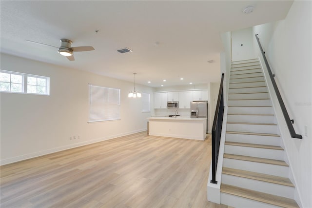 unfurnished living room with light wood finished floors, visible vents, stairway, baseboards, and ceiling fan with notable chandelier