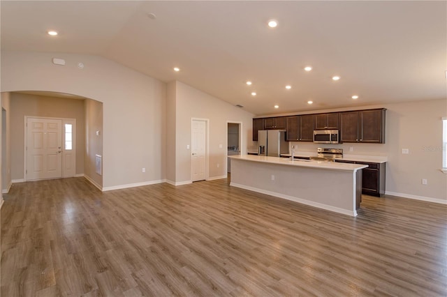 kitchen featuring appliances with stainless steel finishes, a kitchen island with sink, dark brown cabinets, light hardwood / wood-style floors, and vaulted ceiling