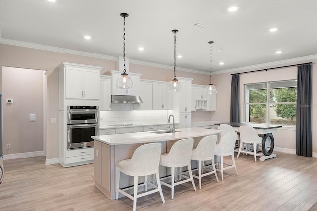 kitchen featuring white cabinets, under cabinet range hood, light countertops, a center island with sink, and double oven