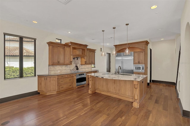 kitchen featuring dark wood finished floors, decorative backsplash, an island with sink, built in appliances, and a sink